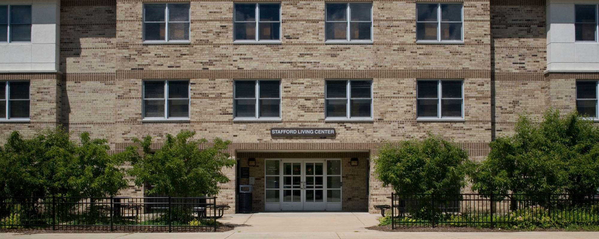 A brick building labeled "Stafford Living Center" has numerous windows and front double doors. Lush green shrubs line the entrance pathway.